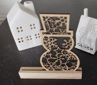 Schneemann Plotterdatei von easy-sticks, kunstvoller Holzschneemann auf einem Holzsockel, zwischen dekorativen Kerzenhaltern. Bringen Sie festliche Stimmung in Ihr Zuhause.