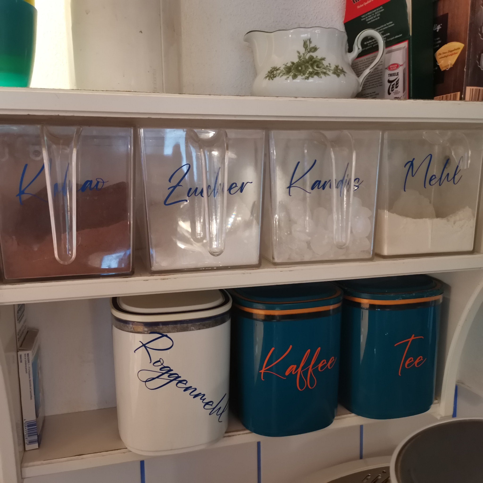 Eine angrenzende Aufbewahrungslösung mit klaren Behältern, die mit Etiketten für Kakao, Zucker, Kandis, Mehl und mehr versehen sind. Perfekt zur Organisation in der Küche. Von easy-sticks, inspiriert von der '138 x Vorratshaltung' Laserdatei.