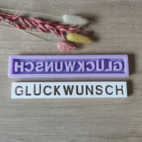Zwei Silikonformen von Easy-Sticks mit dem Schriftzug "GLÜCKWUNSCH", eine lila und eine weiße, dekoriert mit getrockneten Blumen. Perfekt zur Gestaltung individueller Geschenke und Dekorationen.