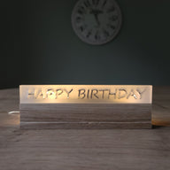 Ein stilvoller Acryl-Schriftzug von Easy-Sticks mit der Aufschrift 'HAPPY BIRTHDAY', sanft beleuchtet und auf einem Holzsockel platziert, ideal für Geburtstagsfeiern.