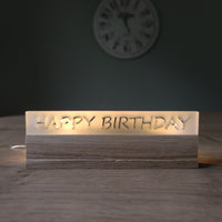 Ein stilvoller Acryl-Schriftzug von Easy-Sticks mit der Aufschrift 'HAPPY BIRTHDAY', sanft beleuchtet und auf einem Holzsockel platziert, ideal für Geburtstagsfeiern.
