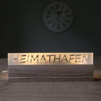 Beleuchtetes Acrylschild 'HEIMATHAFEN' von Easy-Sticks, elegant auf einem Tisch mit Holzoberfläche platziert, ideal als häusliche Dekoration oder Geschenk.