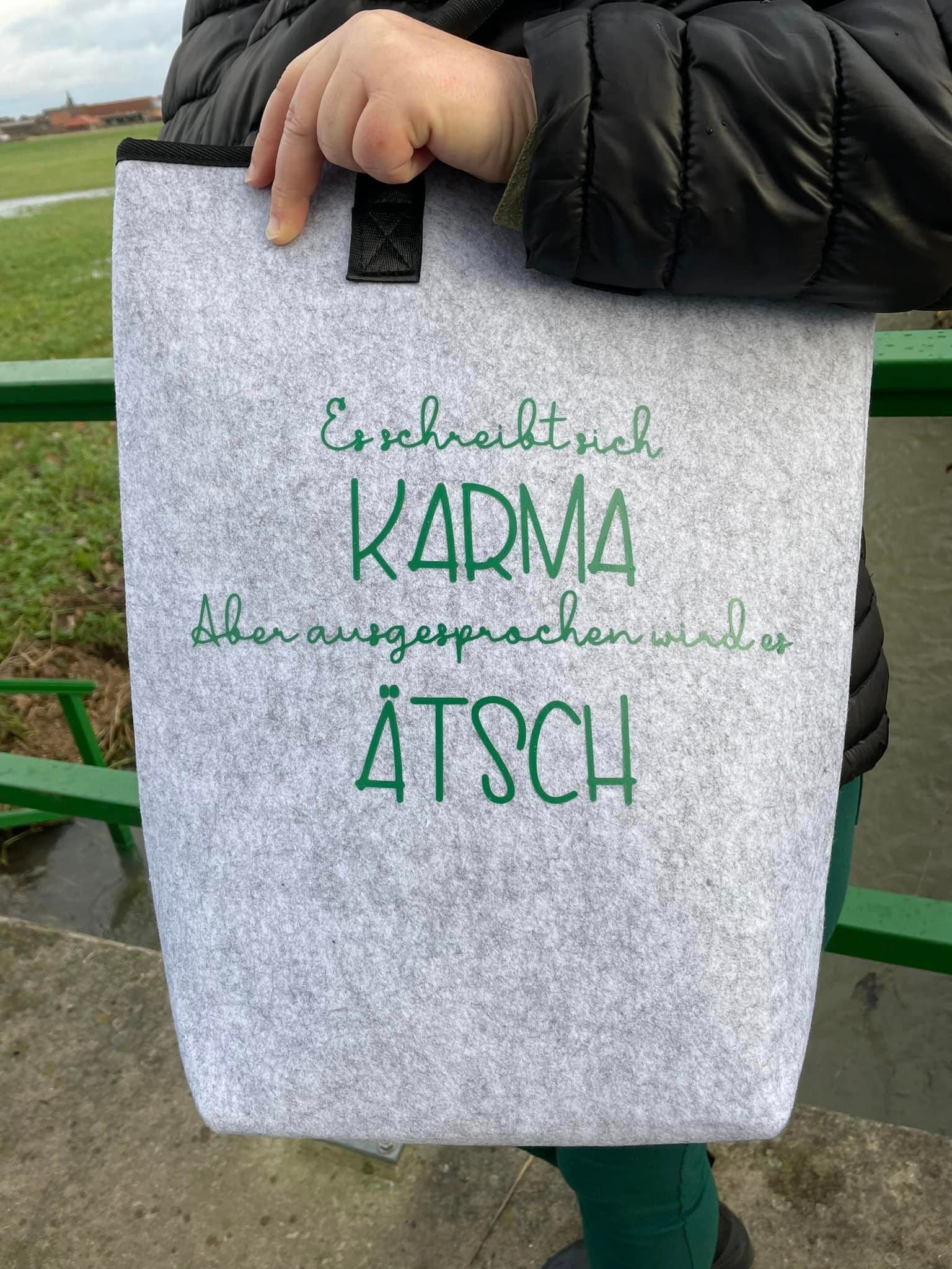 Eine graue Filztasche mit dem Aufdruck "Es schreibt sich Karma Aber ausgesprochen wird es Ätsch" von easy-sticks, perfekt für umweltfreundliche Aufbewahrungslösungen.