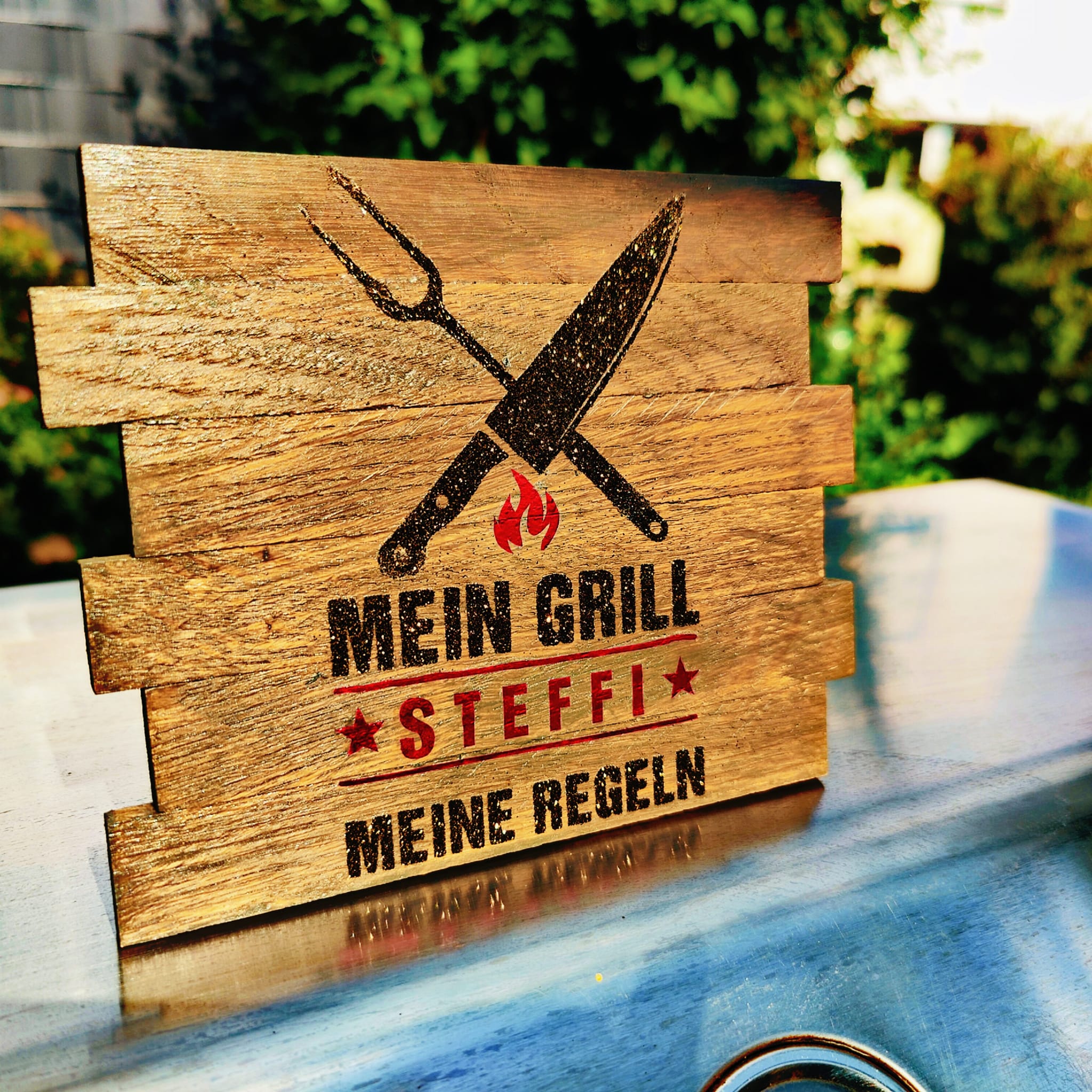 Ein rustikales Holzschild, auf dem steht "MEIN GRILL STEFFI MEINE REGELN", eingerahmt von Grillwerkzeugen, eine kreative Gestaltung von easy-sticks aus der "Grill Sprüche" Plotterdatei.