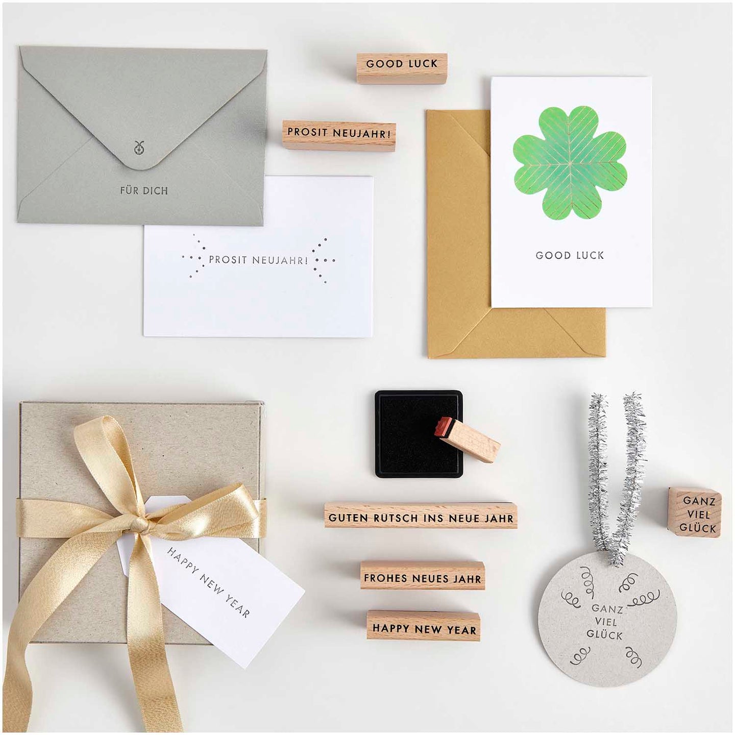 Stempelset Happy New Year FSC 100% von easy-sticks, auf einem weißen Tisch liegend. Das Set umfasst verschiedene Stempel mit Neujahrsgrüßen, eine goldene und eine graue Umschlag sowie eine Geschenkbox mit satiniertem Band und einer Karte, die viel Glück für das neue Jahr wünscht.