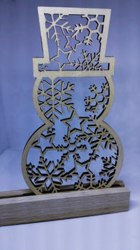 Schneemann Plotterdatei von easy-sticks, in präzisem Holzschnittdesign, steht auf einem Holzgestell, umgeben von winterlichem Flair. Perfekt für Schneidearbeiten in der Adventszeit.
