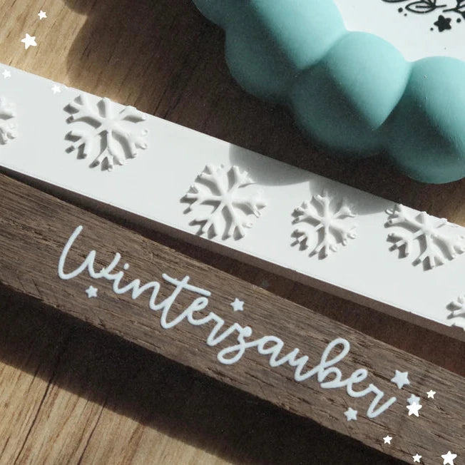 Kreative Nutzung der Easy-Sticks Silikonform - Schneeflocken, dekoriert auf einem Holzbrett mit dem Schriftzug 'Winterzauber' und einer mintgrünen Backform im Hintergrund.