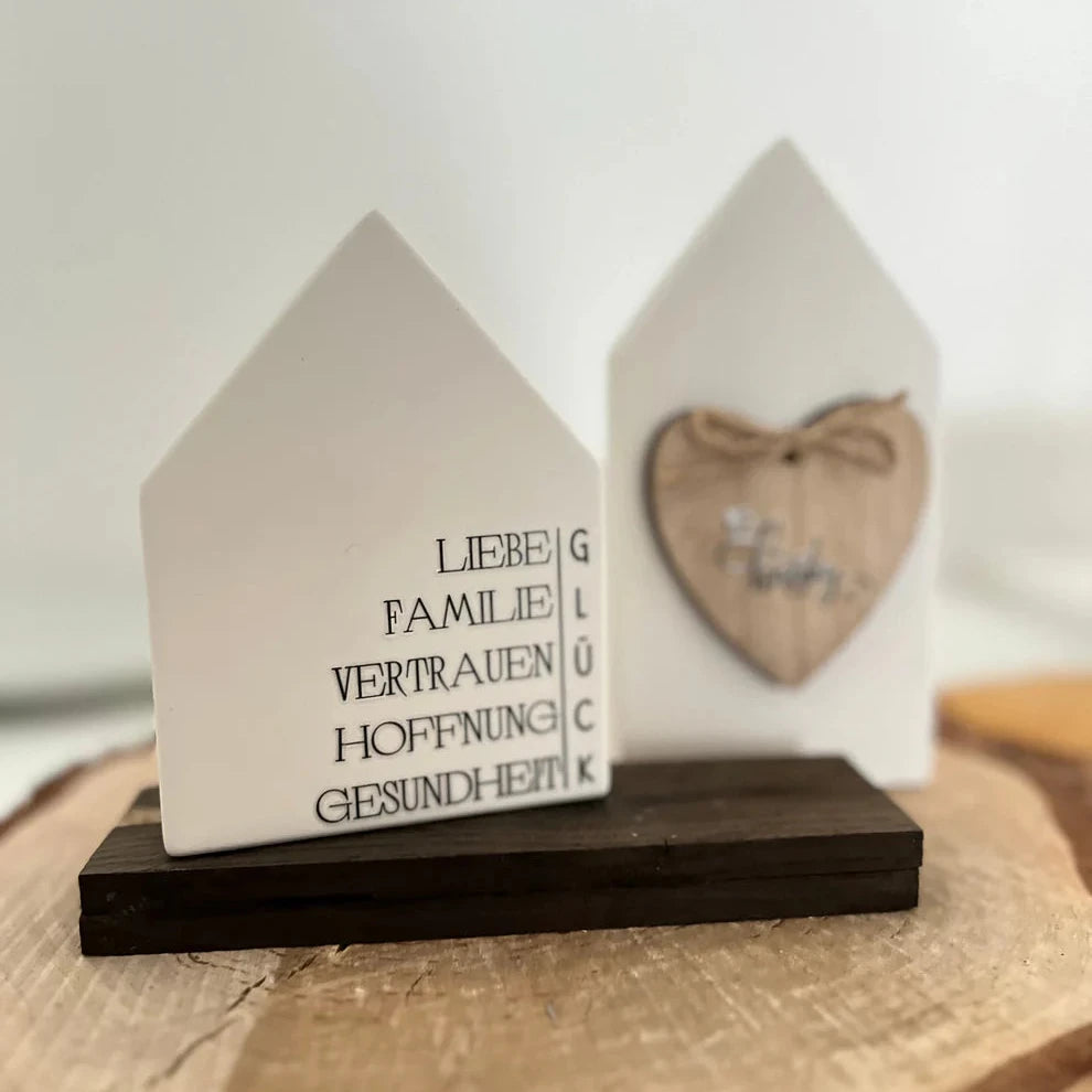 Dekorative weiße Hausfigur mit bedeutenden Wörtern wie 'LIEBE', 'FAMILIE' und 'GLÜCK', kombiniert mit einem herzförmigen Holzanhänger von easy-sticks, Teil der Rub on UV Sticker - randlos Winter A5 Kollektion.