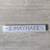 Transluzentes Acrylschild mit dem Schriftzug "HEIMATHAFEN" von Easy-Sticks, arrangiert auf einer Holzfläche.