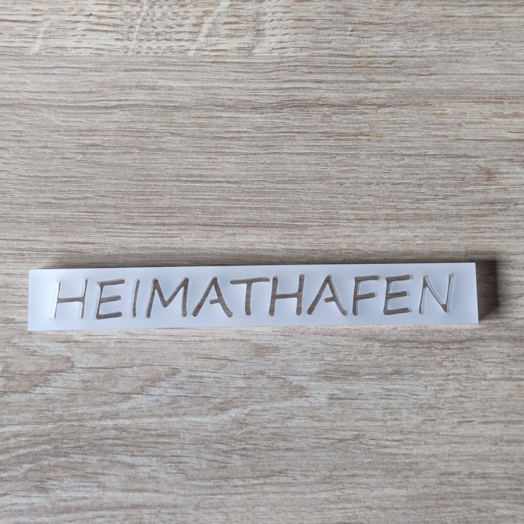 Transluzentes Acrylschild mit dem Schriftzug "HEIMATHAFEN" von Easy-Sticks, arrangiert auf einer Holzfläche.