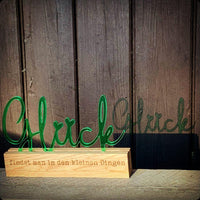 Easy-Sticks Acryl Aufsteller 'Glück' in Grün, elegant platziert auf einer Holzunterlage, mit Schattenwurf an einer Holzwand, ideal für stilvolle Dekoration zu Hause.
