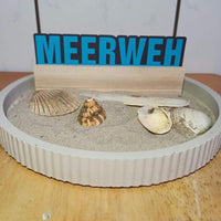 Das Wort 'Meerweh' auf einem sandfarbenen Hintergrund mit Muscheln, passendes Design von easy-sticks.