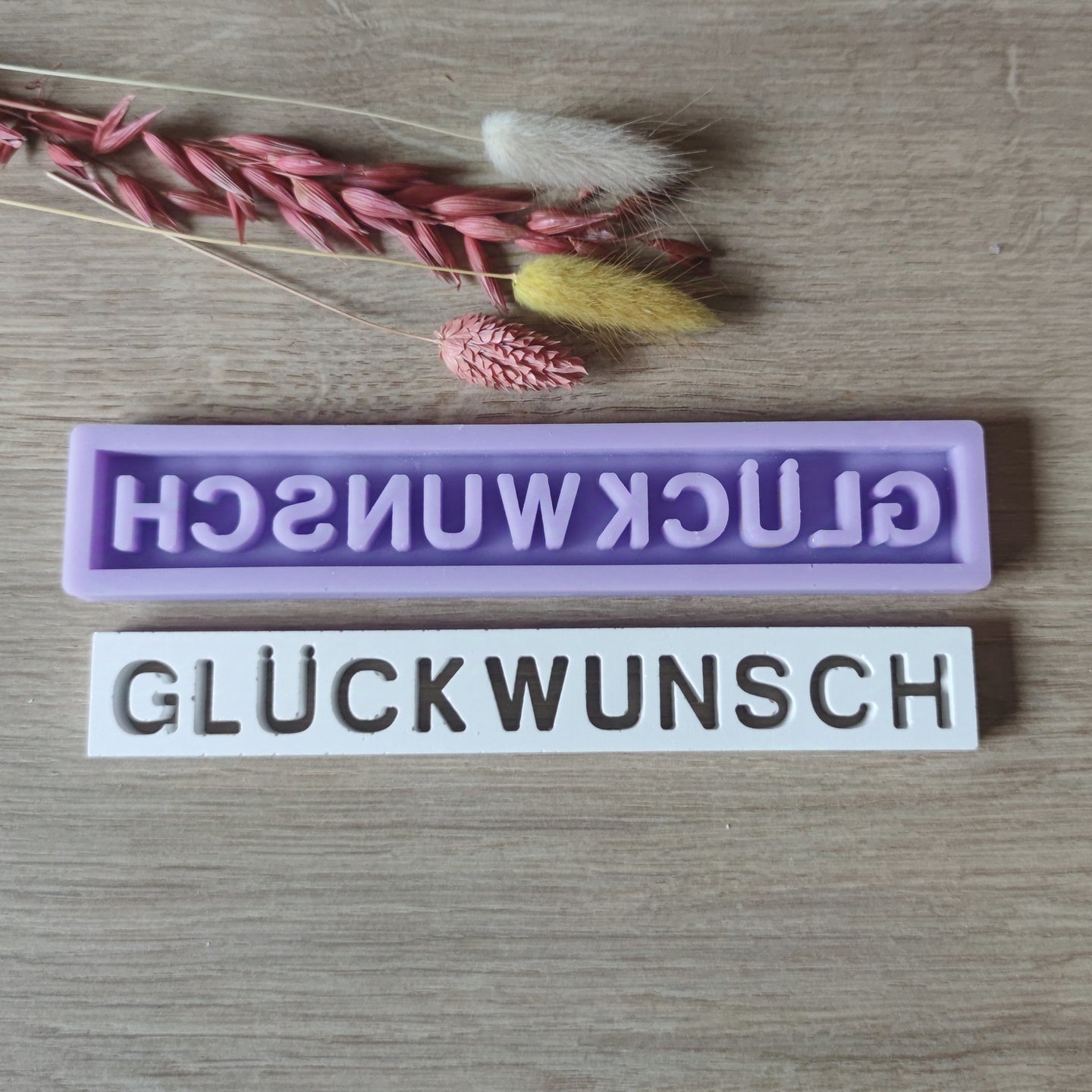Zwei Silikonformen von Easy-Sticks mit dem Schriftzug "GLÜCKWUNSCH", eine lila und eine weiße, dekoriert mit getrockneten Blumen. Perfekt zur Gestaltung individueller Geschenke und Dekorationen.