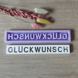 Zwei Silikonformen von Easy-Sticks mit dem Schriftzug "GLÜCKWUNSCH", eine lila und eine weiße, dekoriert mit getrockneten Blumen. Perfekt zur Gestaltung individueller Geschenke und Dekorationen.