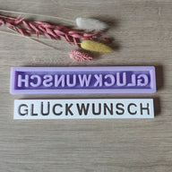 Zwei Silikonformen von Easy-Sticks mit dem Schriftzug "GLÜCKWUNSCH", eine lila und eine weiße, dekoriert mit getrockneten Blumen. Perfekt zur Gestaltung individueller Geschenke und Dekorationen.