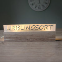 Beleuchtetes Acrylschild mit der Inschrift "LIEBLINGSORT" von Easy-Sticks, stilvoll auf einem Holzsockel angeordnet.