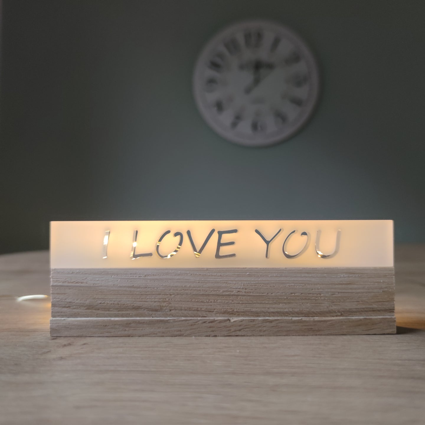 Stylisches Acrylklötzchen 'I LOVE YOU' von Easy-Sticks auf einem Holztisch, elegant beleuchtet im Hintergrund mit einer Wanduhr.
