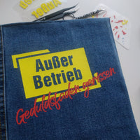 Anwendung der "Außer Betrieb - Geduldsfaden gerissen" Laserdatei von easy-sticks auf einer Jeans mit gelbem Schild und rotem Schriftzug.