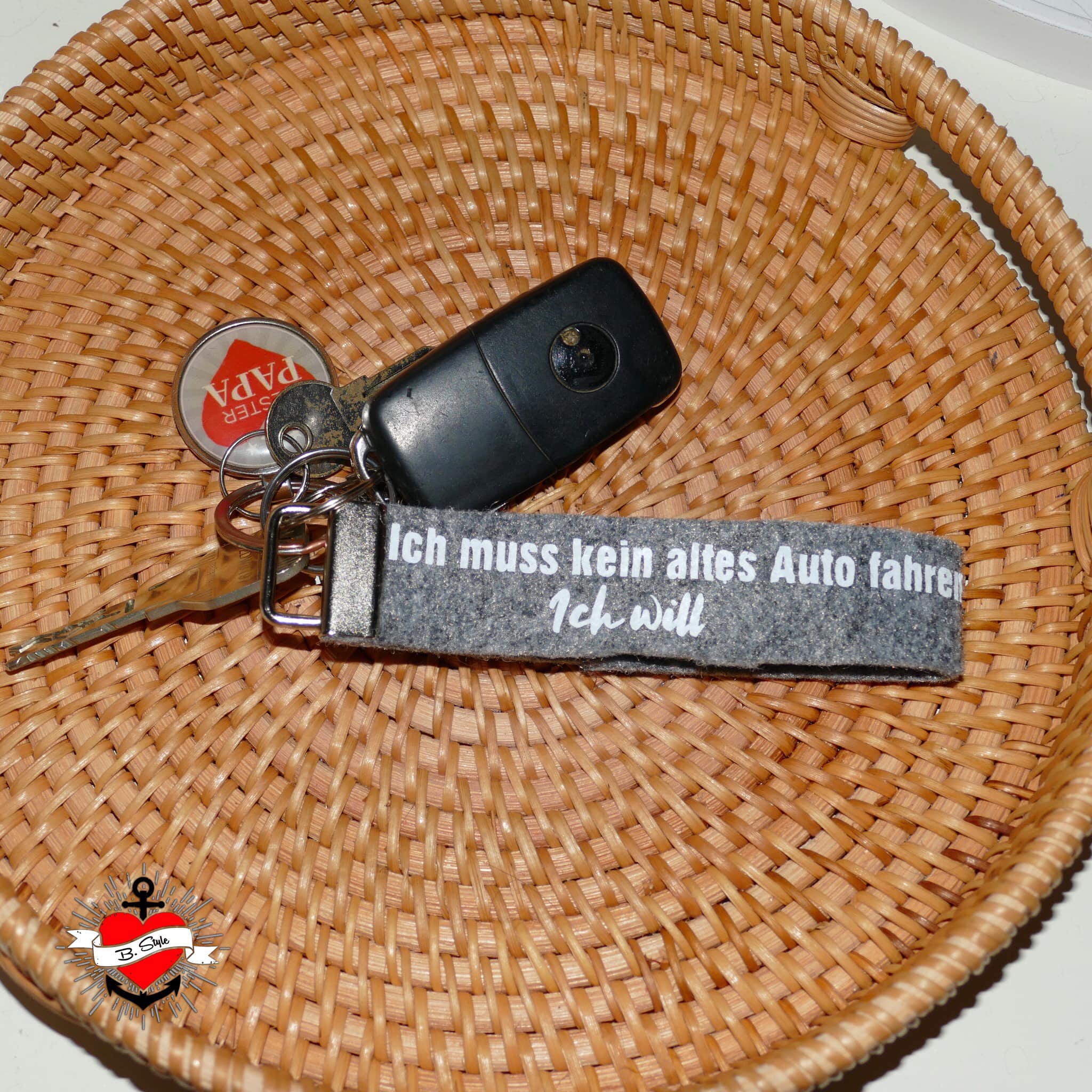 Ein Schlüsselbund liegt in einem Korb mit einem Schlüsselanhänger von easy-sticks, der den Spruch "Ich muss kein altes Auto fahren, Ich will" trägt. Auto Sprüche Plotterdatei.