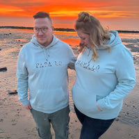Paar steht am Strand bei Sonnenuntergang, beide tragen Hoodies mit den Aufdrucken "PAPA 1.0" und "MAMA 1.0". Das Foto strahlt Freude und Zusammengehörigkeit aus und eignet sich hervorragend für kreative DIY-Projekte mit der Babybauch Plottersprüche Datei von easy-sticks.