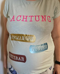 Frau zeigt ein T-Shirt mit den Aufdrucken "ACHTUNG", "SCHWANGER", "HUNGRIG" und "REIZBAR". Das humorvolle Design ist ideal für schwangere Frauen und passt perfekt zu kreativen Ideen mit der Babybauch Plottersprüche Datei von easy-sticks.