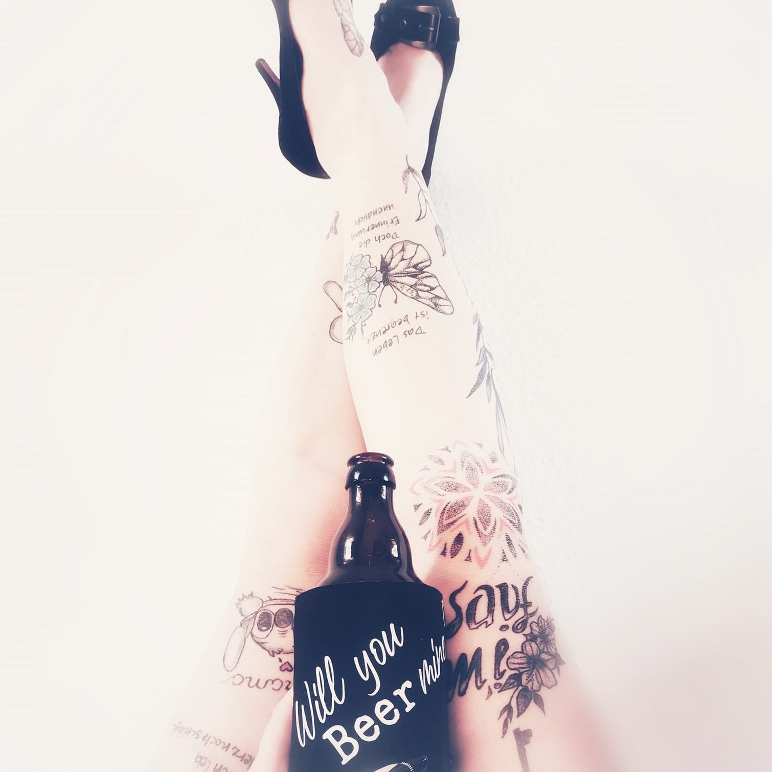 Feminines Bild mit tätowierten Beinen und einem Bier in der Hand, verziert mit einem lustigen Bierwärmer. Ausdrucksstark und kreativ, inspiriert von der Bier Sprüche Laserdatei von easy-sticks.