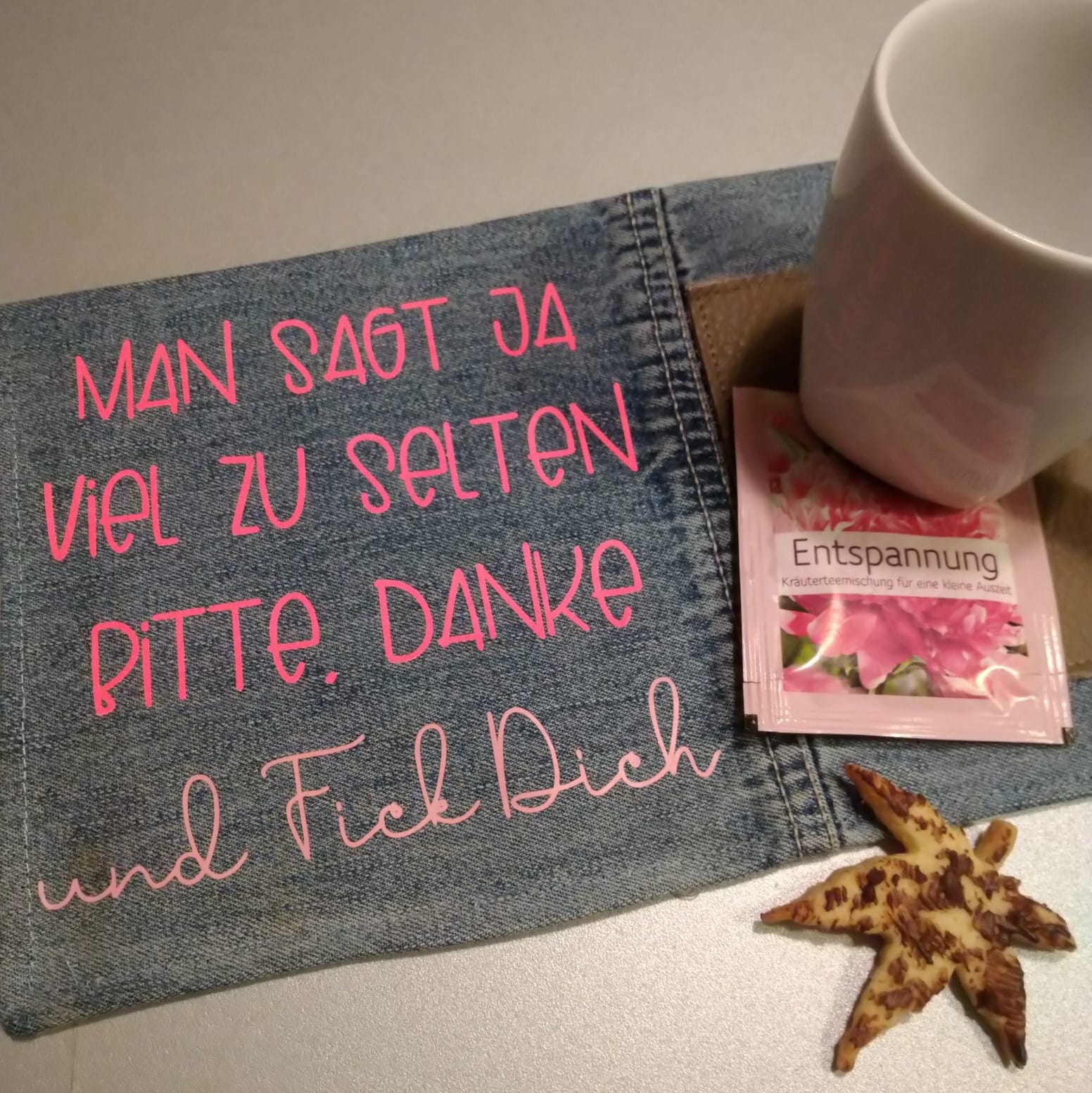 Denim-Stoff mit leuchtend pinker Schrift "MAN SAGT JA VIEL ZU SELTEN BITTE. DANKE. und Fick Dich" darauf liegend. Eine Tasse und ein Teebeutel sind daneben platziert. Entspannende DIY-Idee von easy-sticks.