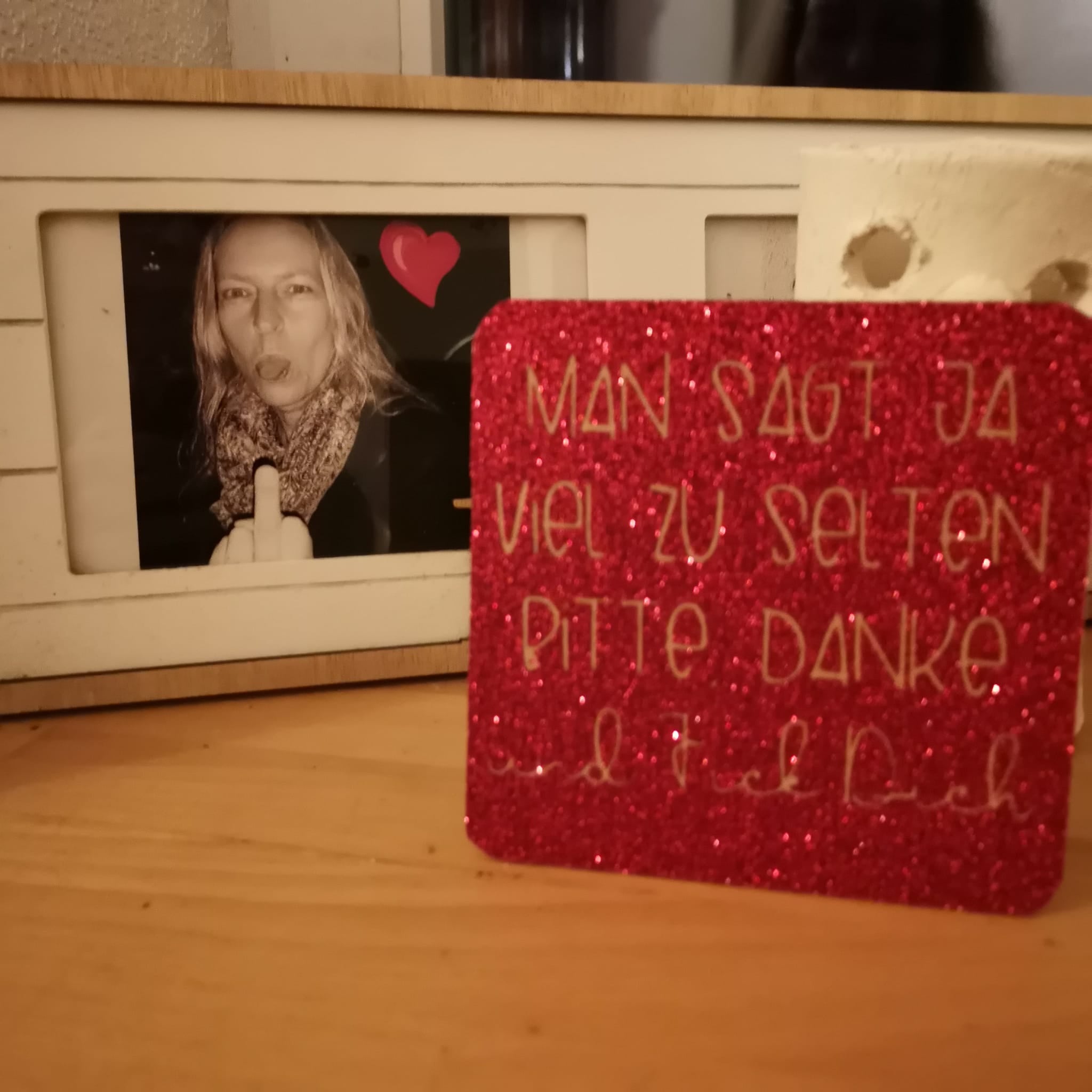 Funkelnde rote Glitzerplatte mit goldener Schrift "MAN SAGT JA VIEL ZU SELTEN BITTE. DANKE. und Fick Dich" im Vordergrund, während ein Foto im Hintergrund sichtbar ist. Kreative home Decor Idee von easy-sticks.