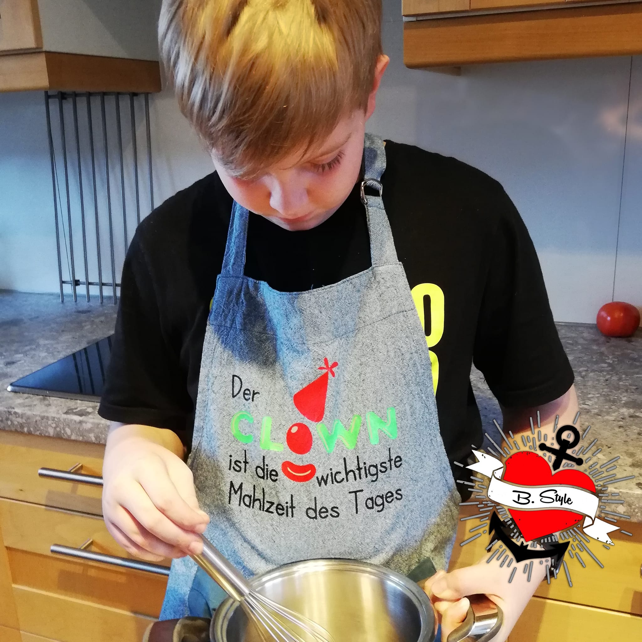 Ein Kind in einer Schürze mit dem Spruch "Der CLOWN ist die wichtigiste Mahlzeit des Tages" von easy-sticks. Entdecke kreative Kochprojekte mit der Clown Laserdatei.