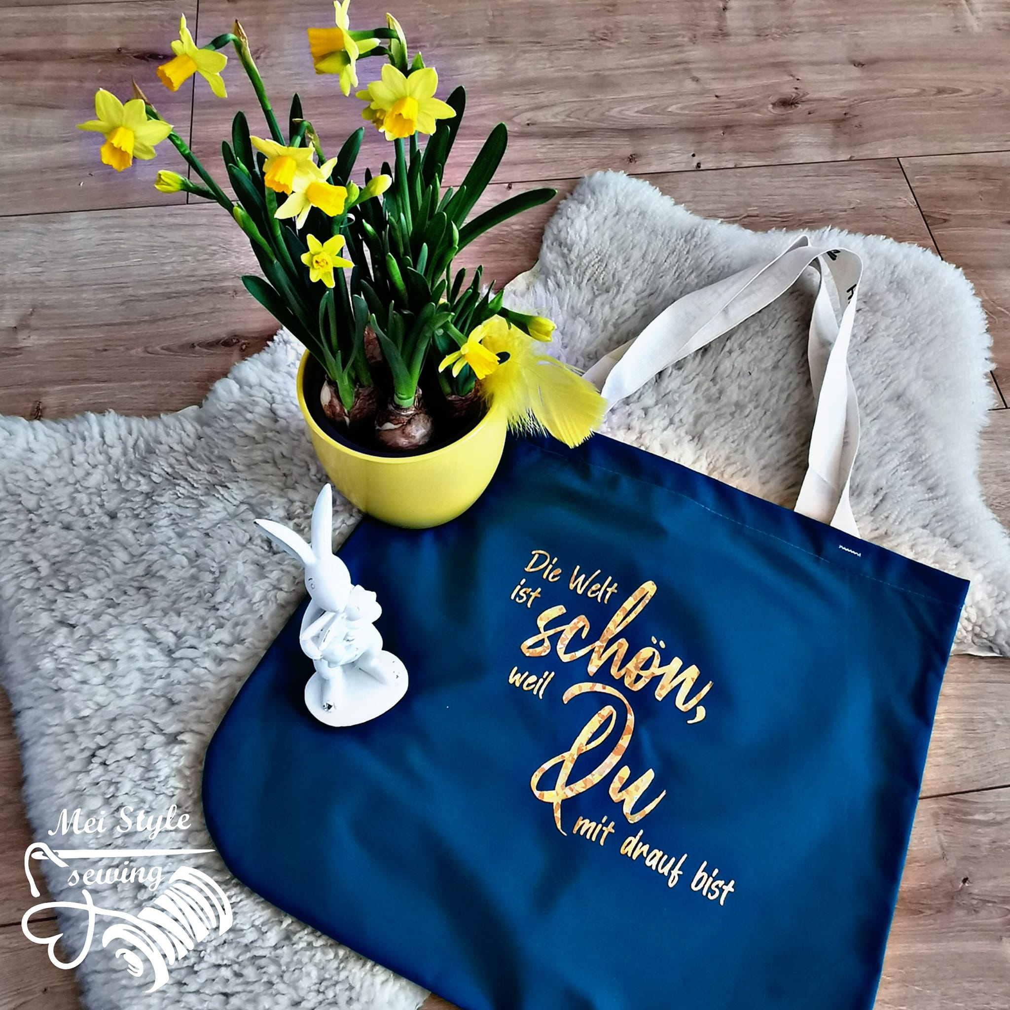 Praktische Tragetasche mit der Aufschrift "Die Welt ist schön, weil Du drauf bist" von easy-sticks, umgeben von fröhlichen gelben Blumen und einem dekorativen Hasen.