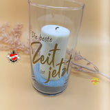 Ein elegantes Glas mit dem Schriftzug "Die beste Zeit ist jetzt" von easy-sticks, dekoriert mit einer weißen Kerze und buntem Sand. Perfekt für kreative DIY-Projekte.
