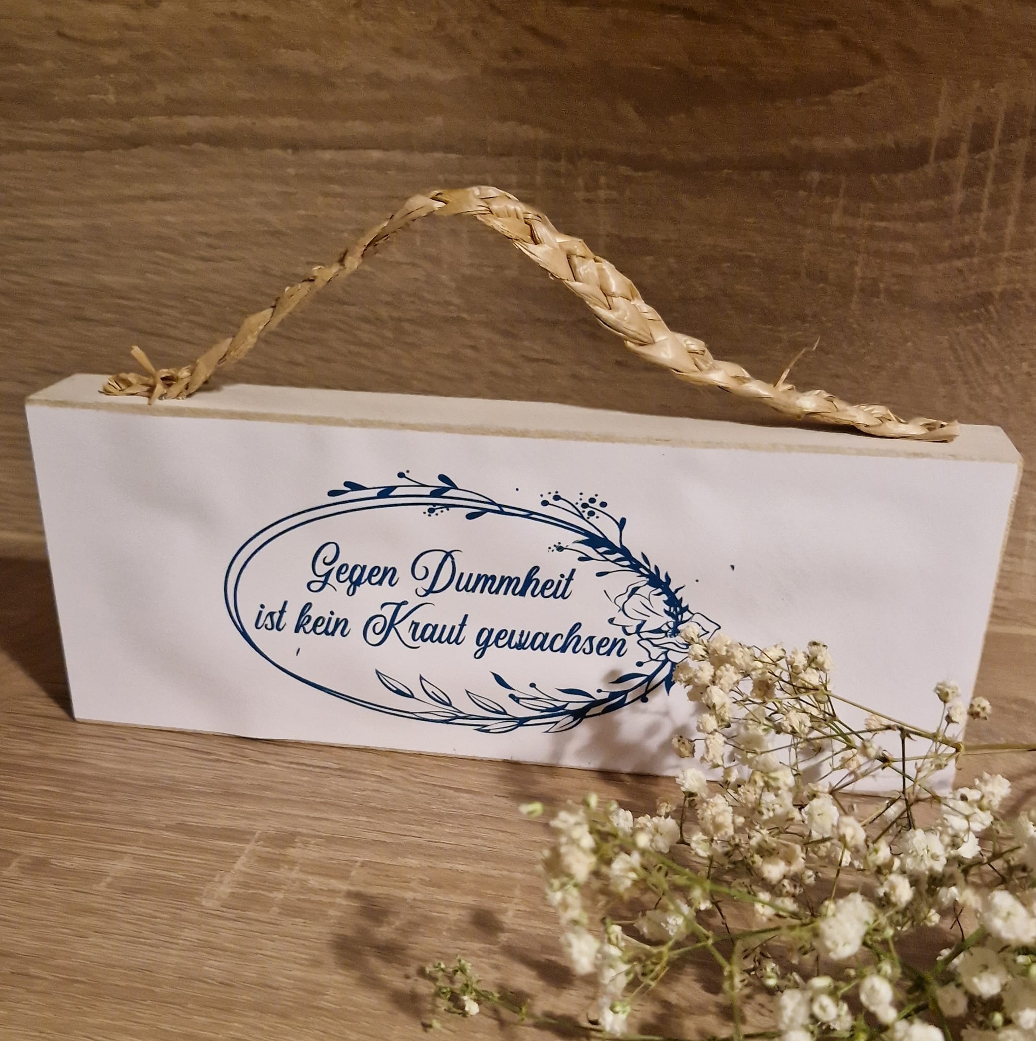 Deko-Schild mit der Aufschrift 'Gegen Dummheit ist kein Kraut gewachsen' von easy-sticks. Ein rustikales Design ergänzt durch eine natürliche Schnur und florale Elemente für kreative Projekte.