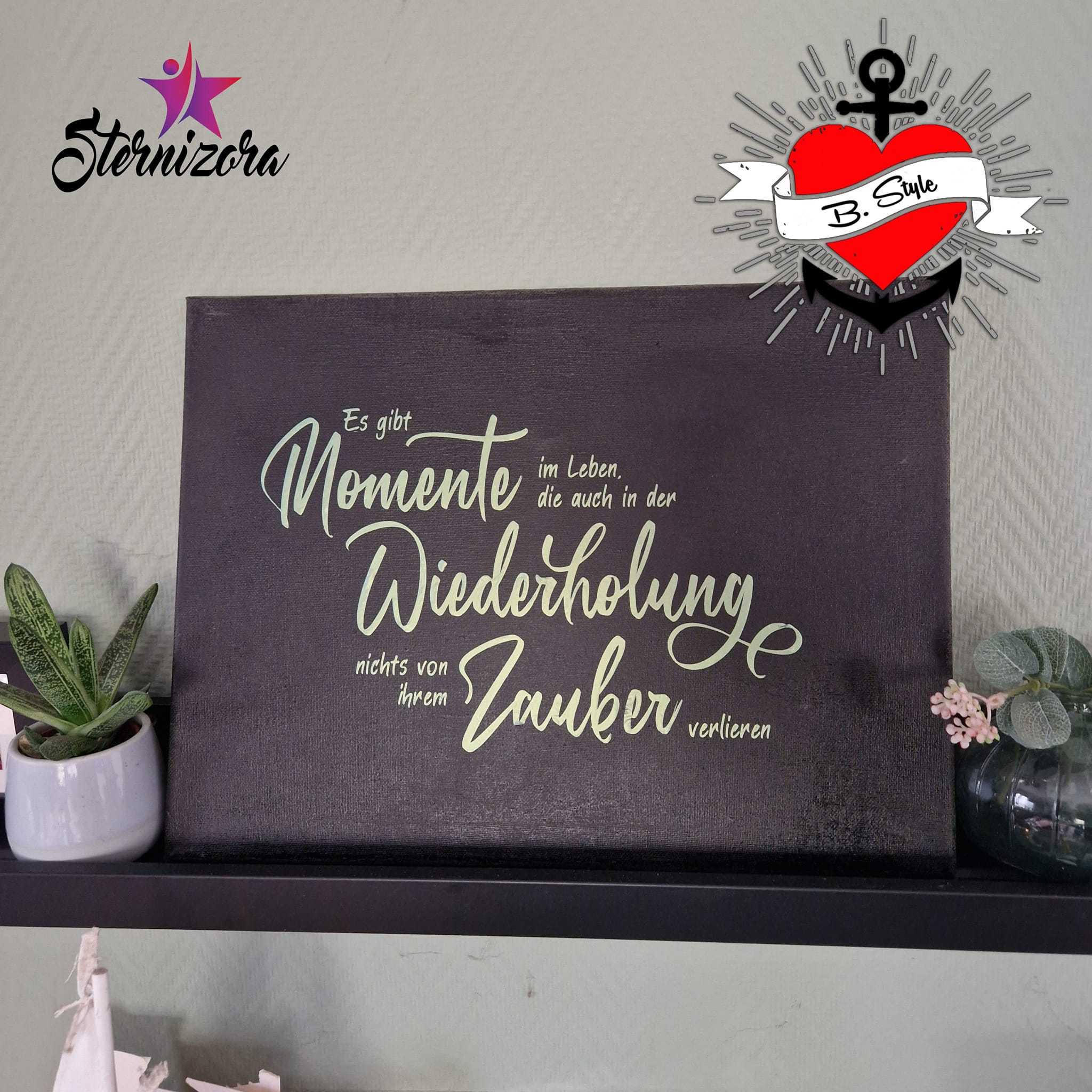 Wandbild mit dem Spruch "Es gibt Momente im Leben die auch in der Wiederholung nichts von ihrem Zauber verlieren" von easy-sticks, präsentiert auf einem Regal neben einer dekorativen Pflanze.