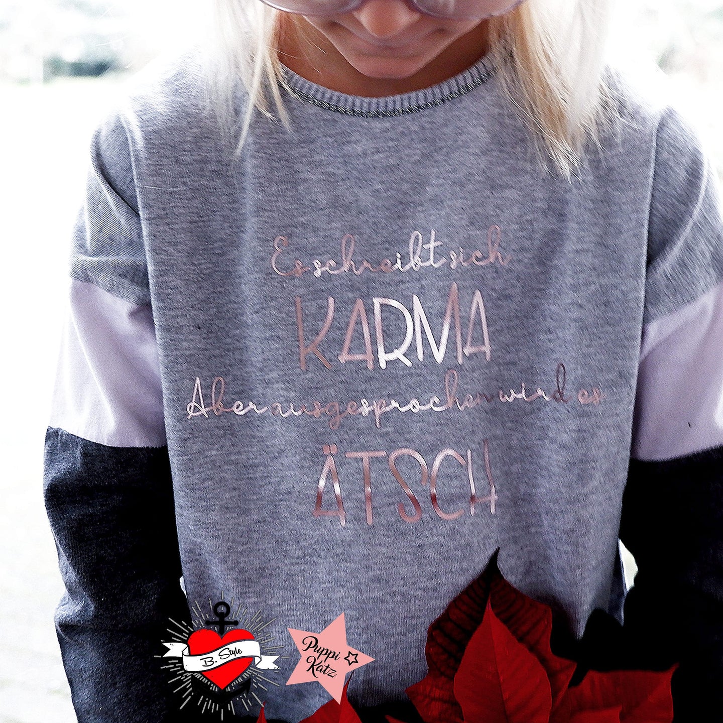 Ein Mädchen trägt ein graues Sweatshirt mit dem Aufdruck "Es schreibt sich Karma Aber ausgesprochen wird es Ätsch" von easy-sticks, perfekt für kreative DIY-Projekte.