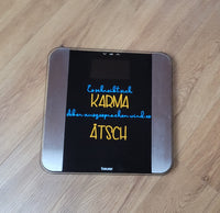 Waage auf dem Boden mit dem Design "Es schreibt sich Karma Aber ausgesprochen wird es Ätsch" in bunten Farben, entworfen mit der Plotterdatei von easy-sticks.