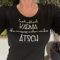 Frau trägt ein schwarzes T-Shirt mit dem Spruch "Es schreibt sich Karma Aber ausgesprochen wird es Ätsch" von easy-sticks, perfekt für entspannte Tageslooks.