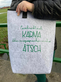 Eine graue Filztasche mit dem Aufdruck "Es schreibt sich Karma Aber ausgesprochen wird es Ätsch" von easy-sticks, perfekt für umweltfreundliche Aufbewahrungslösungen.