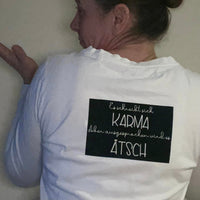 Frau in einem weißen Shirt mit dem Spruch "Es schreibt sich Karma Aber ausgesprochen wird es Ätsch" von easy-sticks, zeigt ihren Rücken, ideal für entspannte Mode.