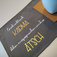 Kreatives Design der Plotterdatei von easy-sticks mit dem Spruch 'Es schreibt sich KARMAB aber ausgesprochen wird es ÄTSCH' auf einem grauen Stoffuntersetzer, ideal für DIY-Projekte.
