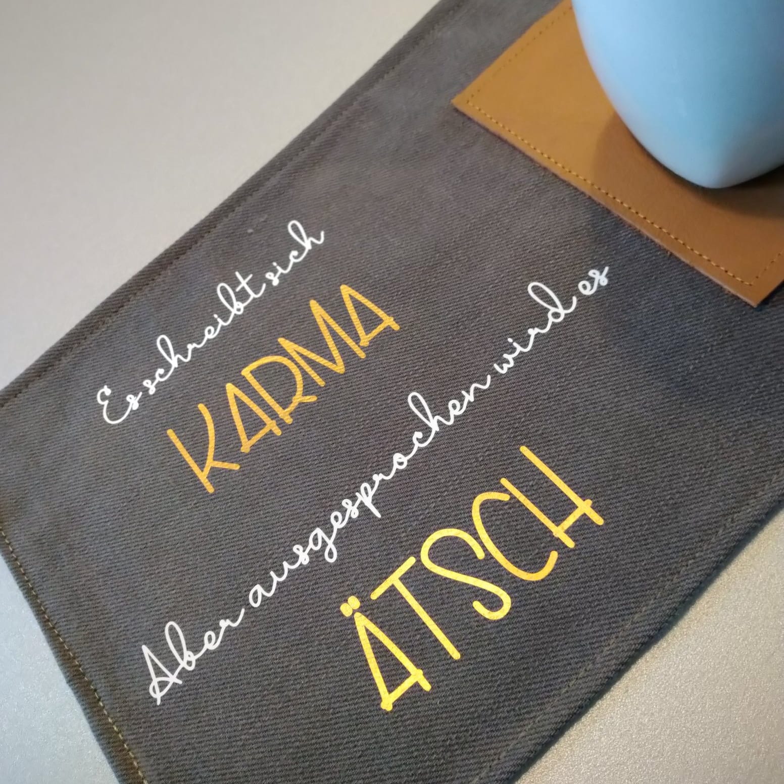 Kreatives Design der Plotterdatei von easy-sticks mit dem Spruch 'Es schreibt sich KARMAB aber ausgesprochen wird es ÄTSCH' auf einem grauen Stoffuntersetzer, ideal für DIY-Projekte.