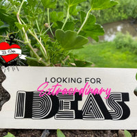 Holzschild mit dem Text 'Looking for extraordinary Ideas' in auffälliger Schrift, umgeben von grünen Pflanzen. Ideal für kreative Projekte mit der Plotterdatei von easy-sticks.