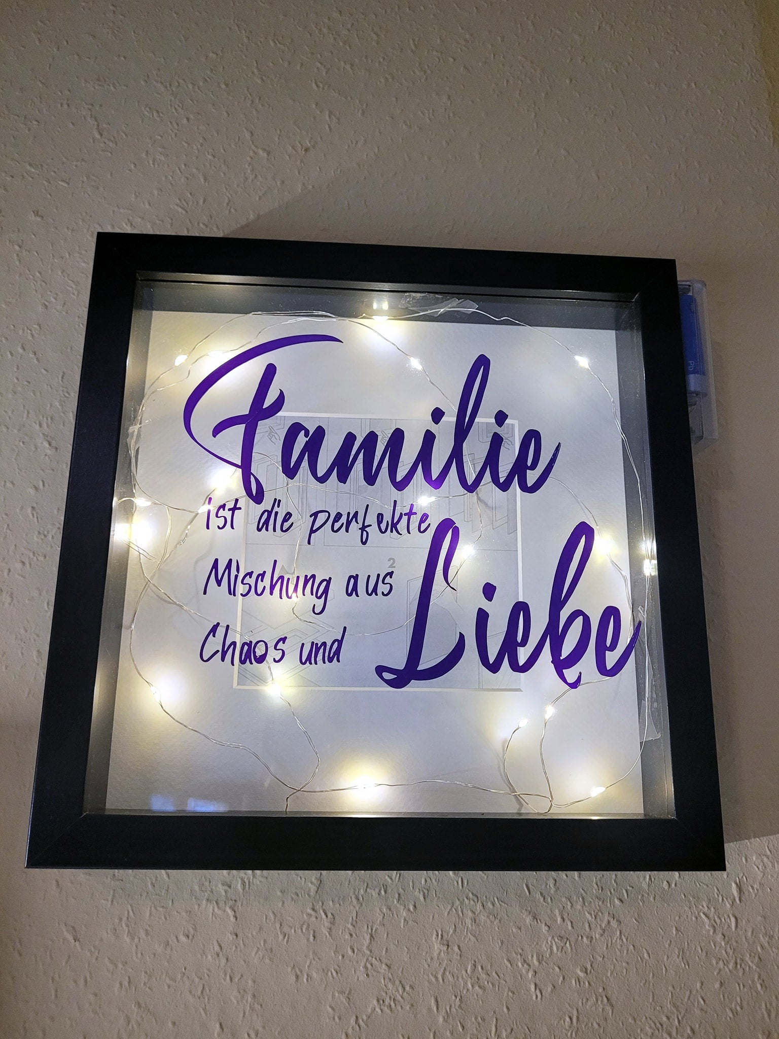 Rahmen mit der Aufschrift "Familie ist die perfekte Mischung aus Liebe und Chaos" von easy-sticks, umgeben von Lichtern, die eine stimmungsvolle Atmosphäre schaffen.