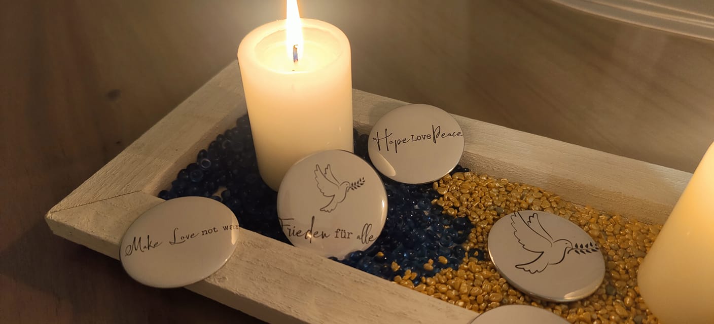 Ästhetische Darstellung einer Friedenskerze aus der Friedenskerze PDF Datei und Plotterdatei von easy-sticks, umgeben von dekorativen Steinen und Badges mit positiven Botschaften wie "Frieden für alle", "Hope Love Peace" und "Make Love not War".
