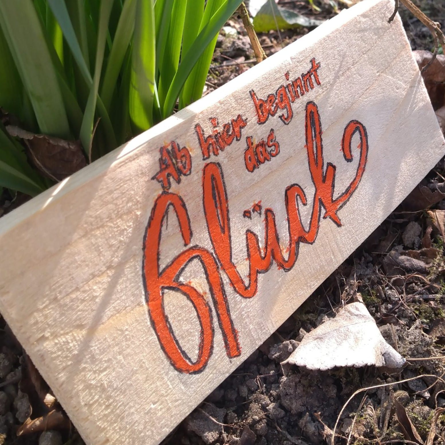 Holzplatte im Freien mit der eingravierten Botschaft 'Ab hier beginnt das Glück', entworfen von easy-sticks. Die leuchtend orangefarbene Schrift setzt einen eindrucksvollen Akzent auf dem natürlichen Holz.