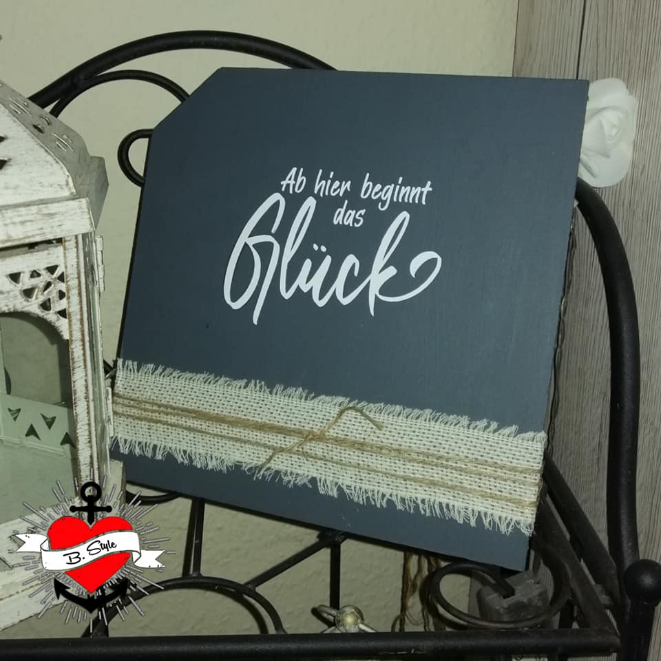 Deko-Schild mit dem Text 'Ab hier beginnt das Glück', gestaltet von easy-sticks. Das Schild zeigt eine elegante Schrift auf einem dunklen Hintergrund, umgeben von dekorativem Material.