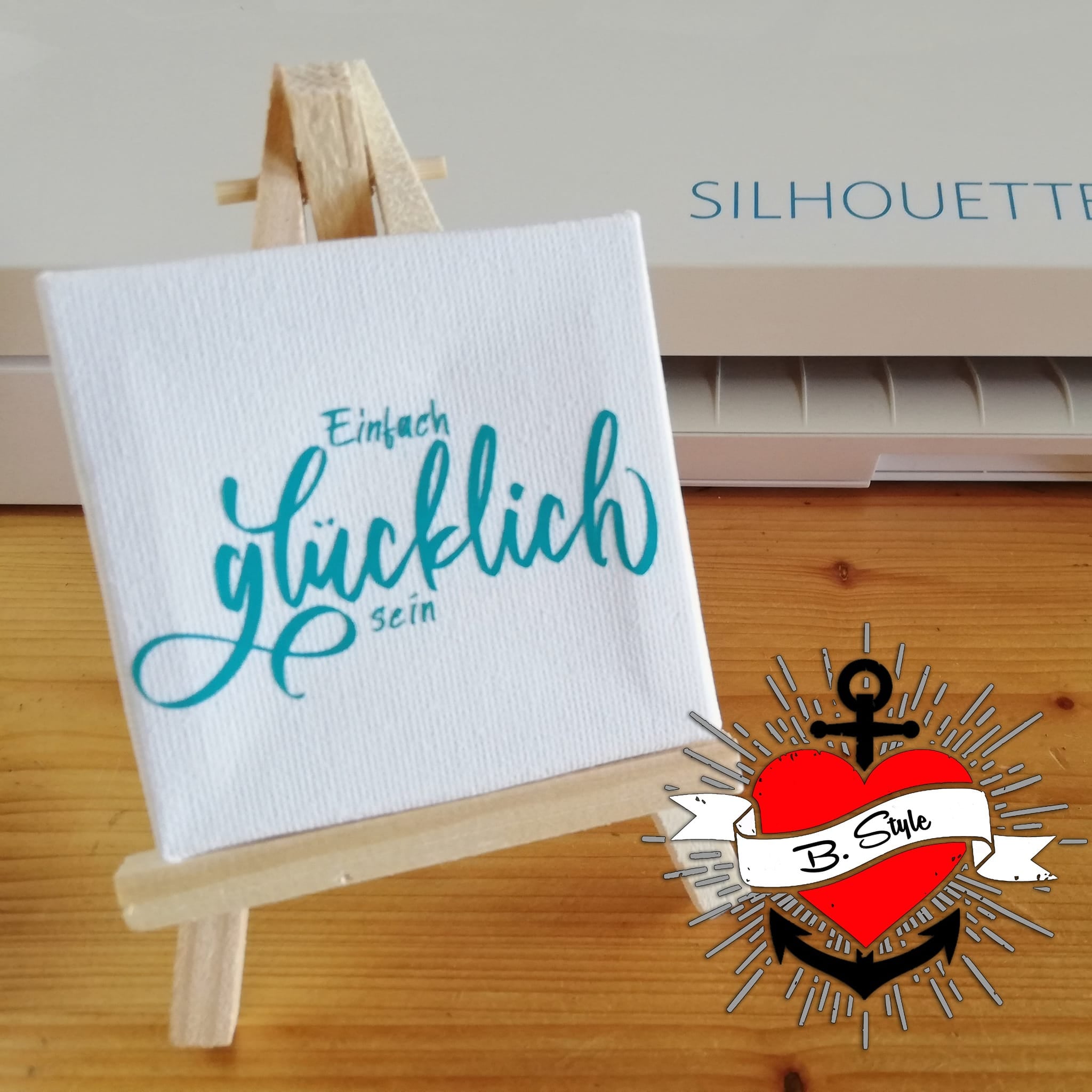 Kreatives Projekt mit der Plotterdatei "Einfach Glücklich sein" von easy-sticks, präsentiert auf einer kleinen Staffelei. Das Design zeigt den inspirierenden Schriftzug "glücklich sein" in stilvollem Türkis und erzeugt eine positive Atmosphäre.