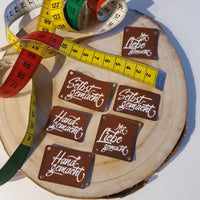 Kreative Grüße Plotterdatei von easy-sticks: Detailansicht von handgemachten Etiketten aus Leder auf einer Holzplatte, dekoriert mit buntem Maßband. Ideal für DIY-Projekte.