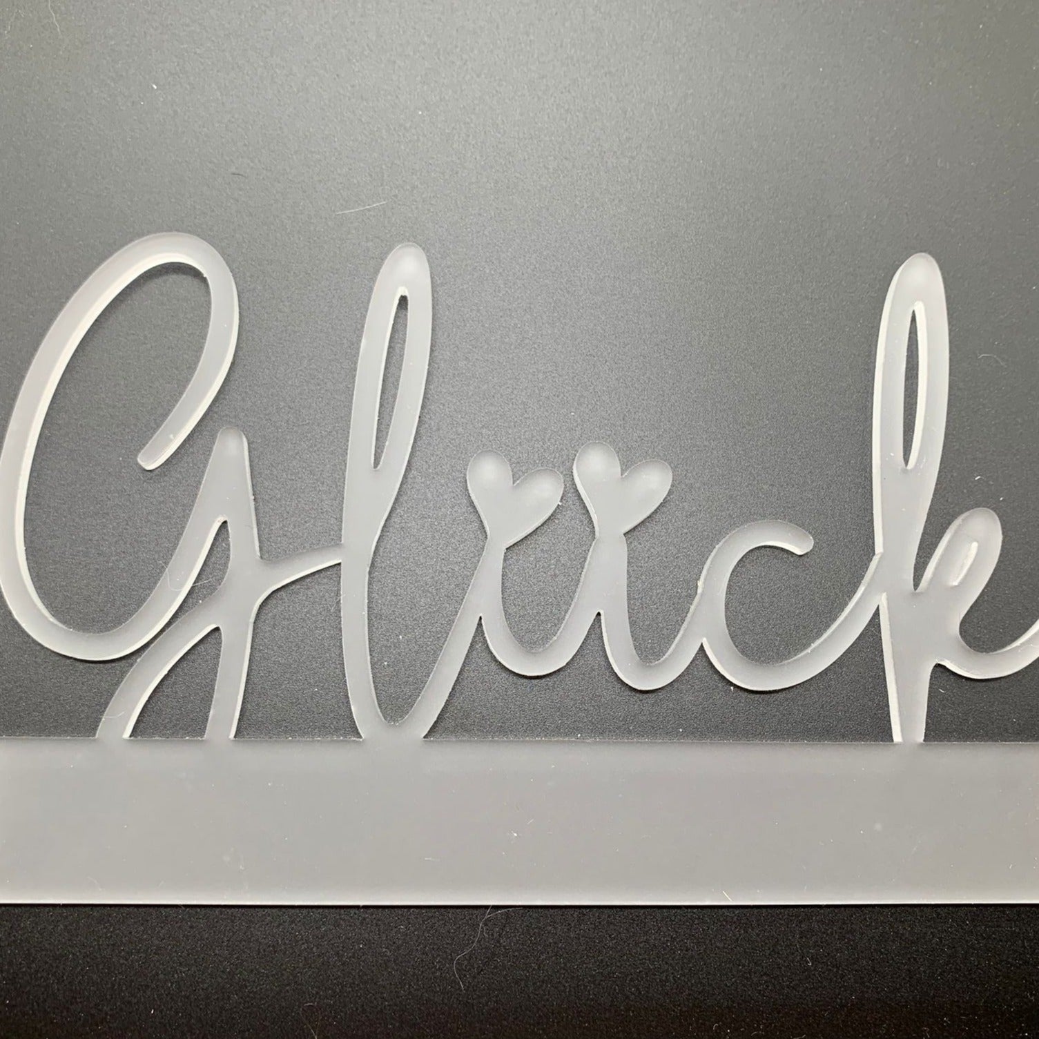 Transparenter Easy-Sticks Acryl Aufsteller 'Glück', vor einem dunklen Hintergrund, hebt sich durch das elegante Design ab, ideal für besondere Geschenkideen.