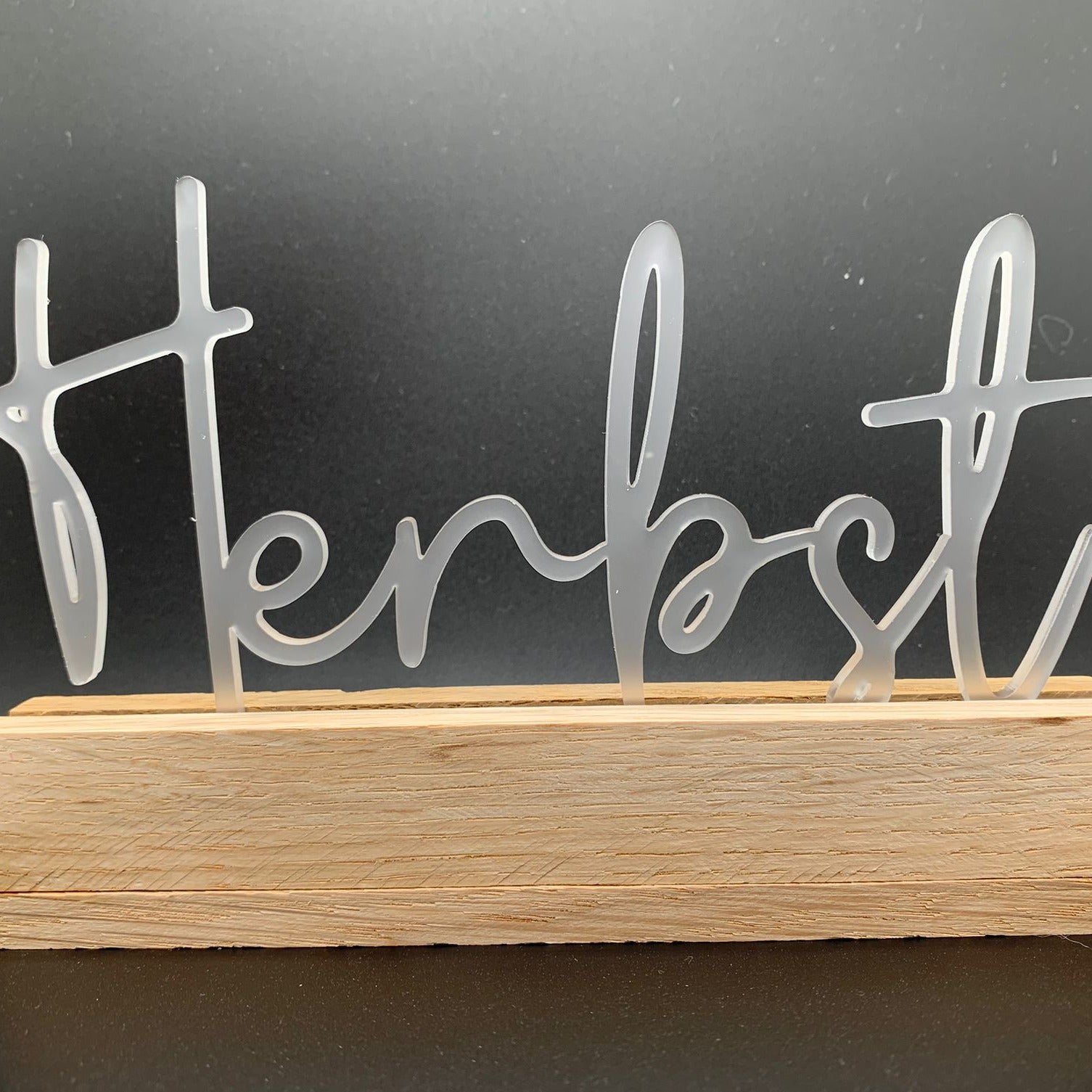 Minimalistischer Herbstschriftzug von Easy-Sticks Acryl Aufsteller auf einem Holzblock, vor einem dunklen Hintergrund.