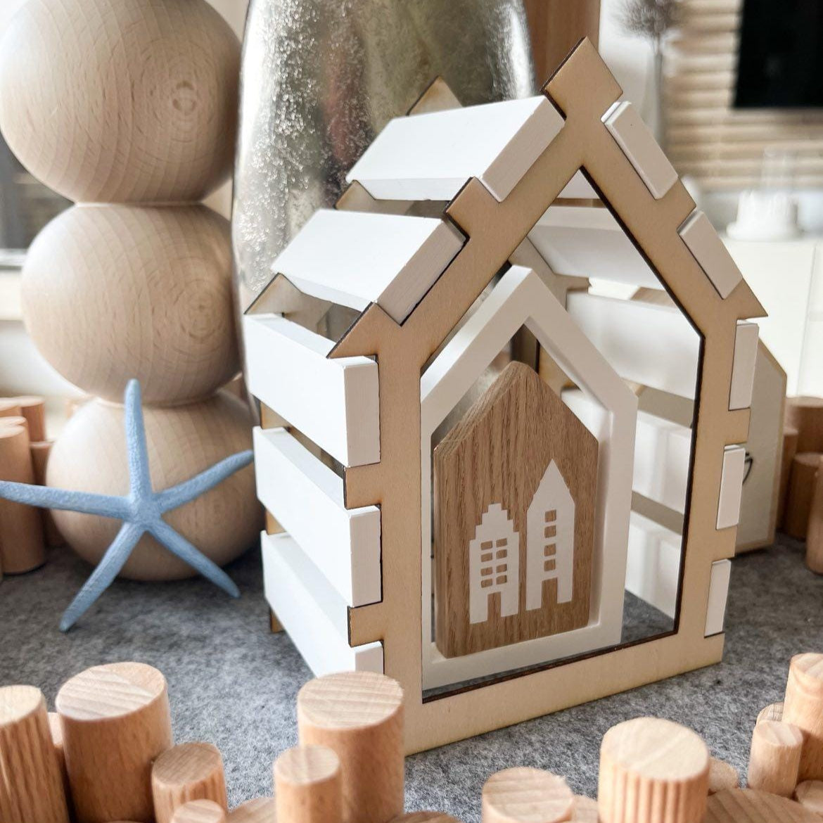 Kreativer Laserdatei Haus Rahmen von Easy-Sticks, dekoratives Dekohaus mit bunten Holzspielzeugen im Hintergrund.
